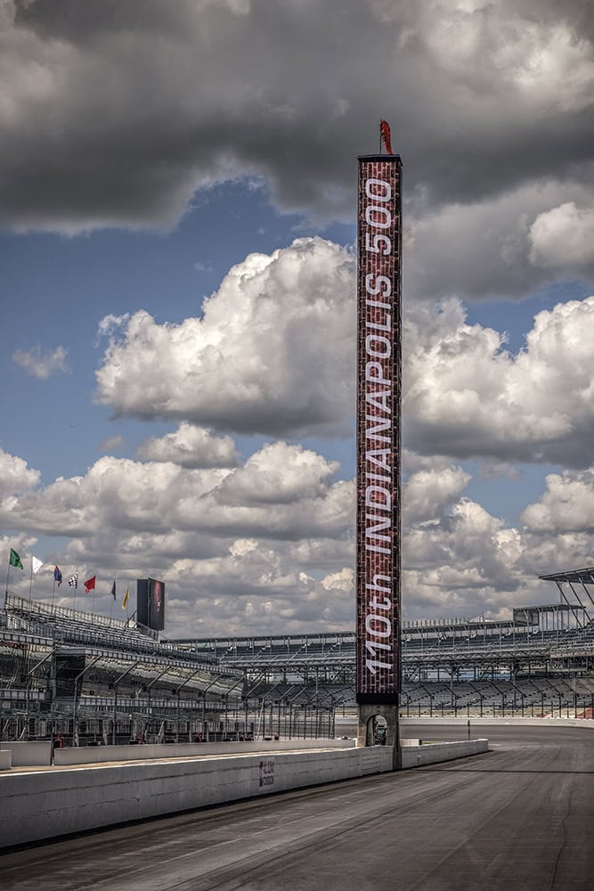The Indianapolis Motor Speedway scoring pylon rises above the empty track and grandstands, with “110th Indianapolis 500” displayed vertically and dramatic clouds filling the sky overhead.