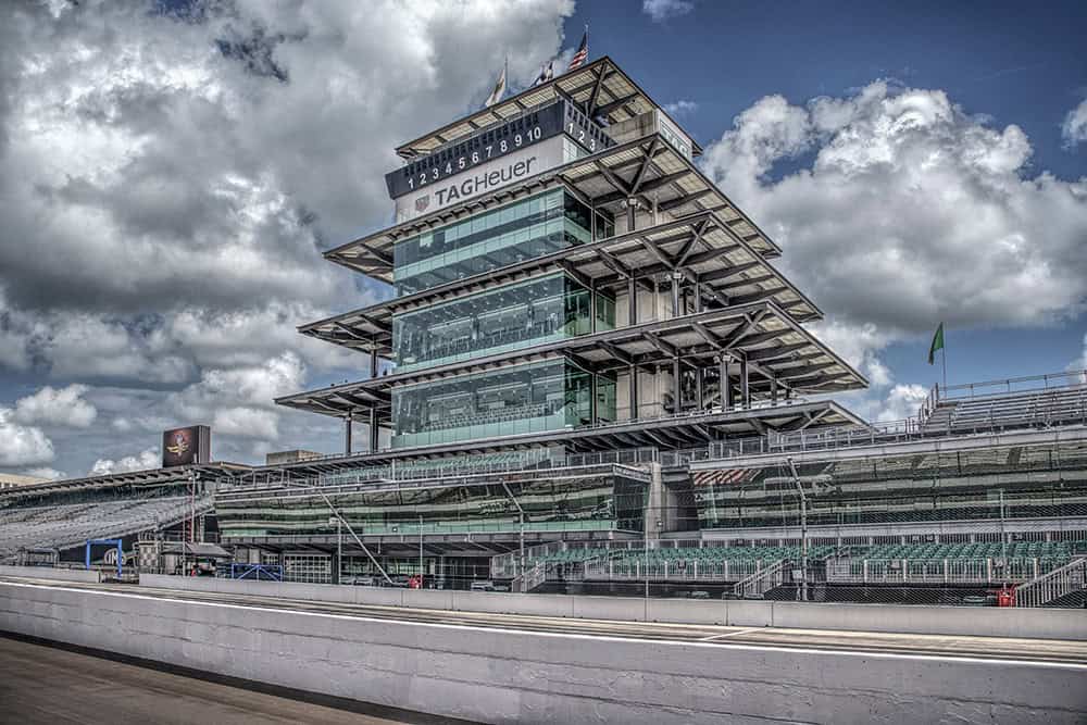The modern pagoda at Indianapolis Motor Speedway rises above the track, grandstands, and fencing under a dramatic sky filled with large white and gray clouds.
